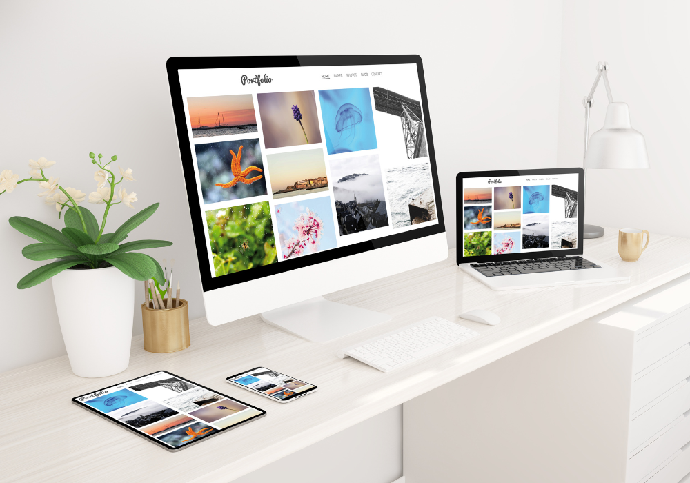 Mobile responsive A modern, minimalistic workspace featuring a desktop computer, laptop, tablet, and smartphone all displaying a photography portfolio website with a clean grid layout. The desk is white and sleek, complemented by a white keyboard and mouse, a small potted plant, a pencil holder, and a cup. A stylish desk lamp is mounted on the wall above the laptop.