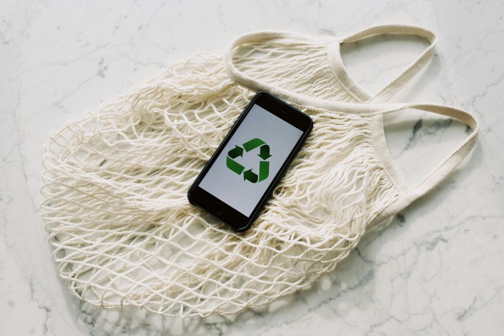 Photo by ready made: https://www.pexels.com/photo/mobile-phone-with-green-recycling-sign-and-mesh-bag-3850512/ A Green recycling logo on a mobile phone screen on top of a reusable bag.