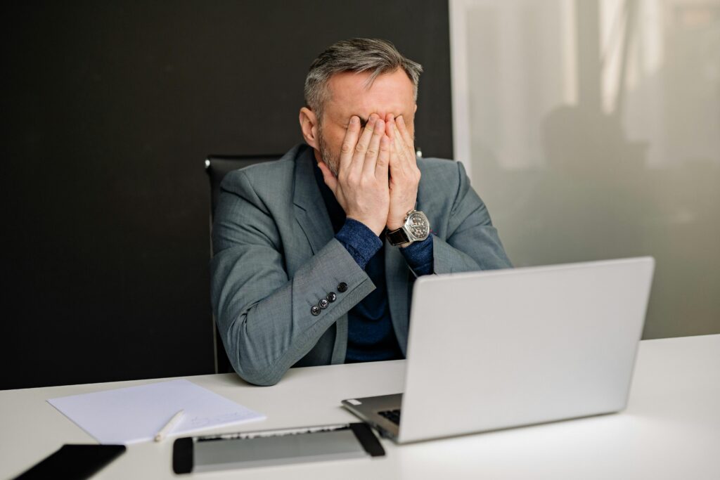 A man in a grey suit holds his head in his hands out of frustration. He is sat behind a laptop.